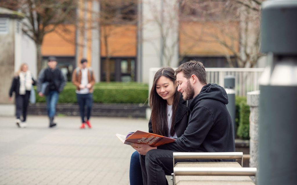 An international student and domestic student sit together on a bench outside of our West Campus and study a textbook together.